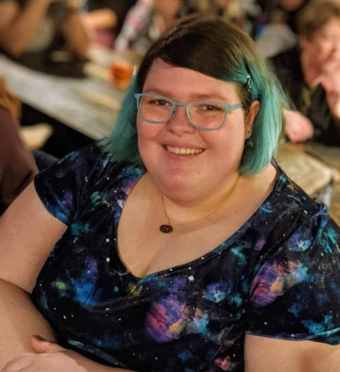 The logo of "Beyond Access" consulting services. It is the International symbol of accessbily shown as a wheelchair user mid push iuin shilloutte shown in blur. It is blue, yellow and pink with a rainbow autism inclusion symbol on the wheel.